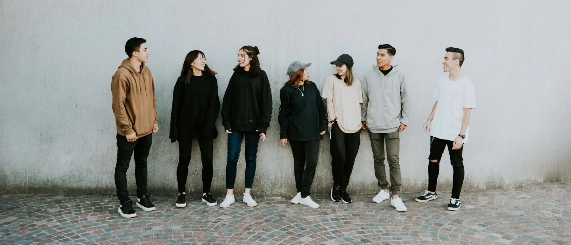 Group of seven young adults standing in a line against a plain wall, engaging in conversation during their visit to the Yo Bro | Yo Girl Youth Initiative, dressed in casual attire.