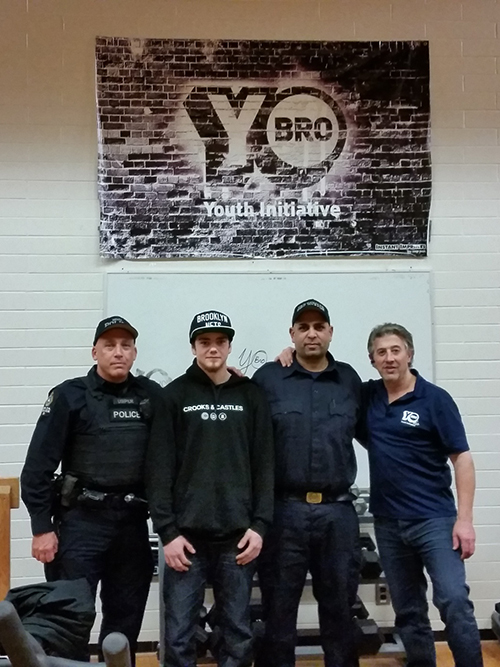 Four men, including two police officers and two civilians, stand smiling before a brick wall with a 