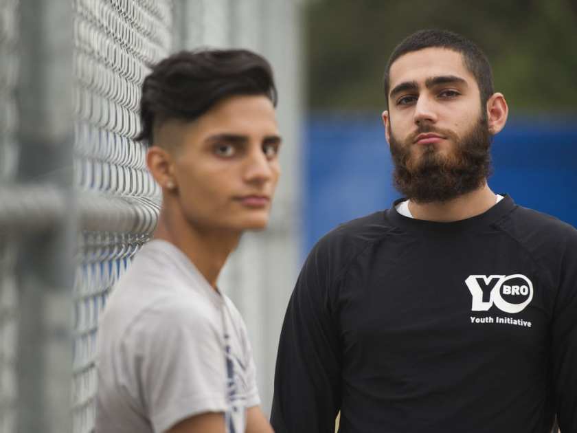 Two young men involved in a gang prevention program standing near a chain-link fence, one in the foreground with a beard and black shirt, the other slightly out of focus in the background.