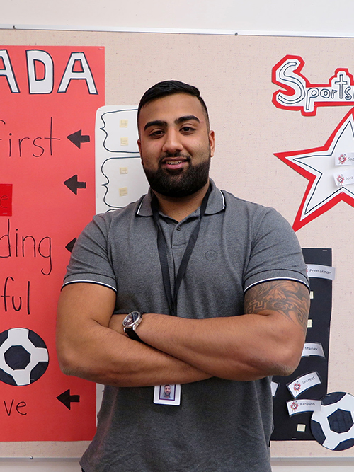 Man with a beard and tattoos, wearing a gray polo and a badge, standing in front of a school bulletin board with various educational posters labeled 